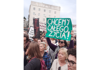 Manifestation avec des personnes sur une place avec une femme à lunettes qui tend une pancarte verte