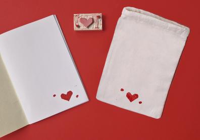un carnet blanc ouvert et une pochette blanche à côté avec un petit coeur rouge dessiné. Au milieu des deux objets il y a une tampon de la forme du coeur