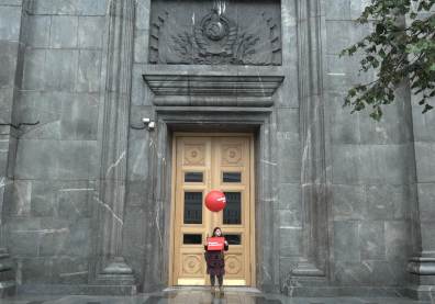 Une jeune femme au pied d'un immeuble gris, devant une porte marron, tenant une pancarte rouge et un ballon rouge