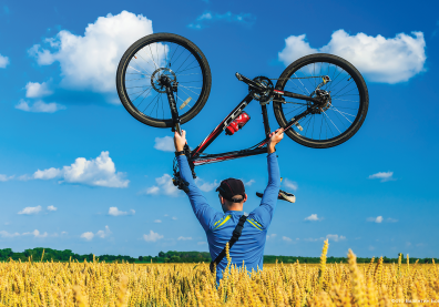 Un homme de dos dans un champ de blé porte un vélo au dessus de sa tête. Le ciel est bleu avec quelques nuages