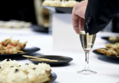 Une table avec une nappe blanche et des plats. Un main attrape une coupe de champagne