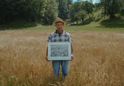 Un homme dans un champ de blé portant un chapeau et une chemise à carreaux. Il tient dans ses mains un cadre avec une photo