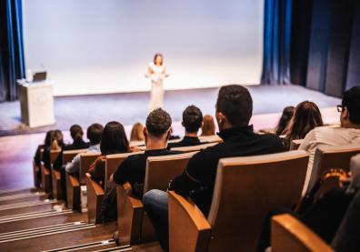 Un amphi avec vue sur l'estrade et le public de dos, une femme donne une conférence
