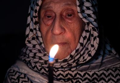 Portrait d'une vieille dame portant un keffieh palestinien blanc dans l'obscurité, tenant une lampe.
