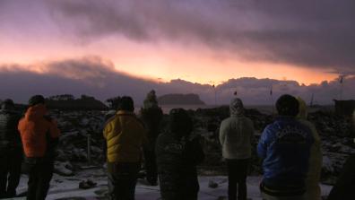 Plusieurs personnes en doudoune et capuches sont de dos et regardent un coucher de soleil face à la mer, le sol est recouvert de neige.