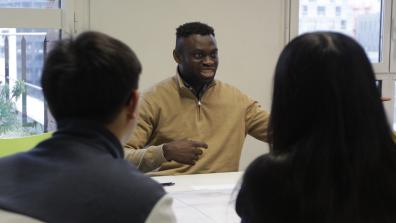 Un homme parle à deux personnes de dos. Ils sont assis autour d'une table dans une salle de classe