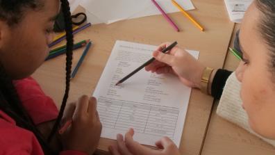 Two students work at a desk and write on a sheet of paper.