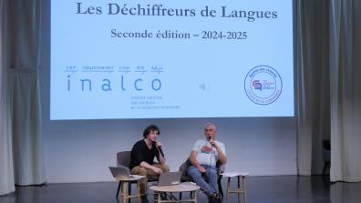 Two men speak into a microphone on the auditorium stage, with a powerpoint behind them.