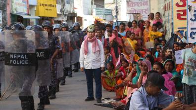 Une rue au Népal. A gauche, la police avec des casques et protections, à droite, des femmes et enfants assis par terre. Au milieu, un homme en chemise porte un foulard et un masque de tigre sur la tête