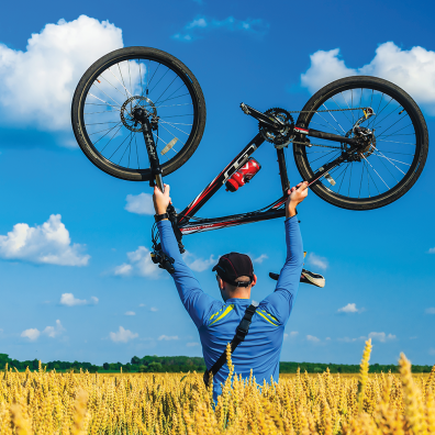 Un homme de dos dans un champ de blé porte un vélo au dessus de sa tête. Le ciel est bleu avec quelques nuages