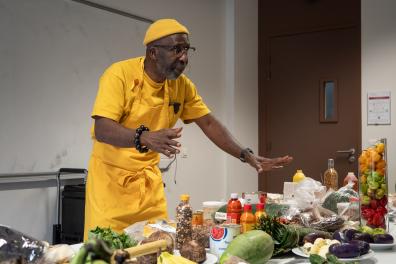 Un chef en tablier parle devant une table garnie de légumes et d'épices