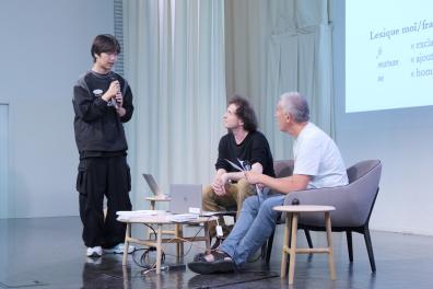 Two men are seated on the auditorium stage, and a student stands in front of them and asks them a question