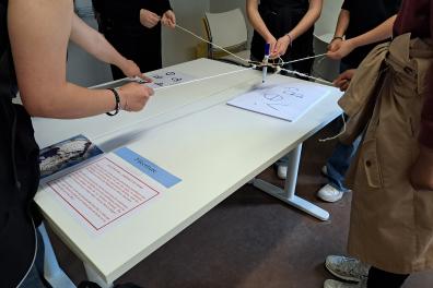 Les élèves se tiennent debout autour du bureau, ils tiennent chacun un morceau de fil. Au milieu de chaque fil se trouve un stylo reliant tous les fils