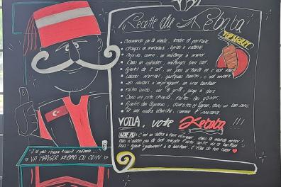 Sur un tableau est dessiné un homme avec une moustache et un chapeau et à sa droite un texte à la craie