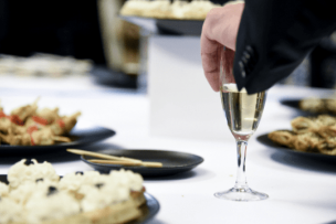 Une table avec une nappe blanche et des plats. Un main attrape une coupe de champagne