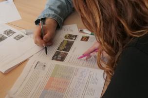 We see the hands of two people on a desk, working on written and illustrated documents.