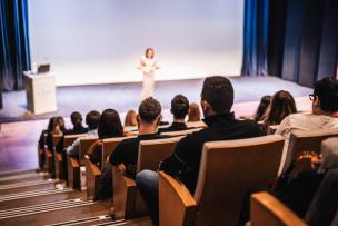 Un amphi avec vue sur l'estrade et le public de dos, une femme donne une conférence