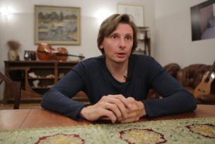 Un homme est assis à une table dans un salon, il y a des instruments de musique derrière lui