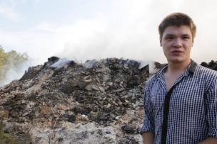 Un jeune homme en chemise se tient en premier plan devant une foret et un tas de gravas entrain de bruler